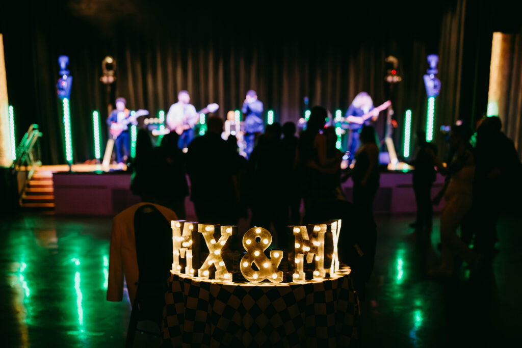 A Mx. and Mx. sign light up as a punk band plays in background at LGBTQ wedding.