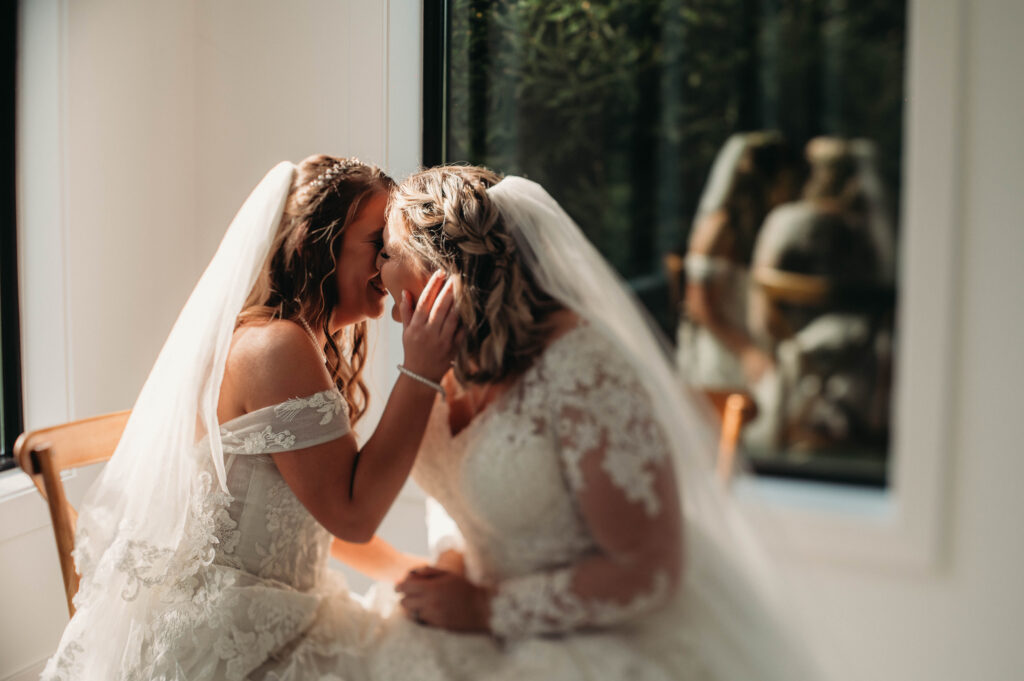 Brides kiss in a sunlit white room on wedding day.