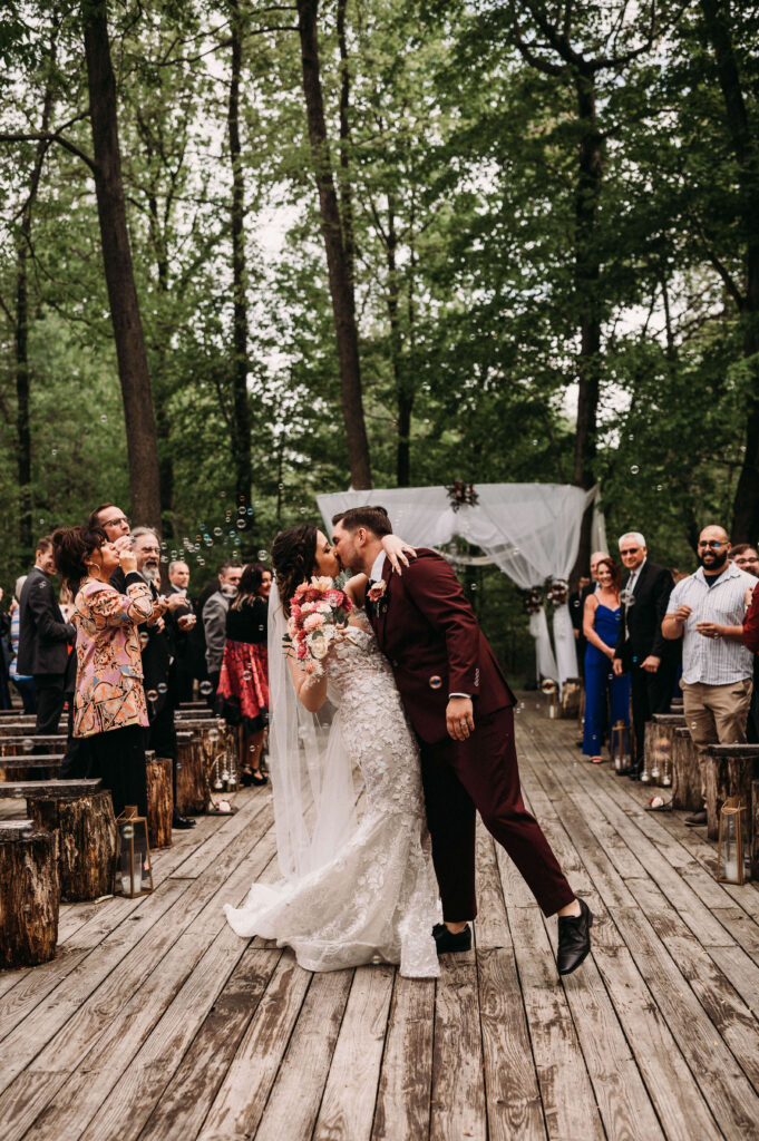 Bride and groom kiss in forest ceremony with bubbles surrounding them.