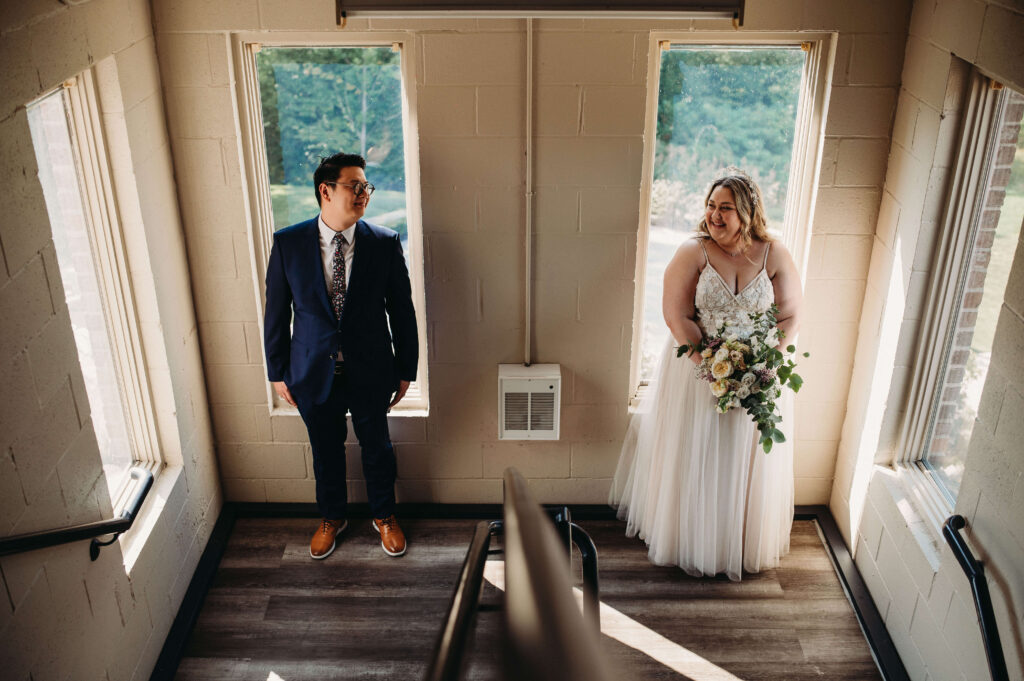 Bride and groom stand in front of windows on sunny day at a unique venue.
