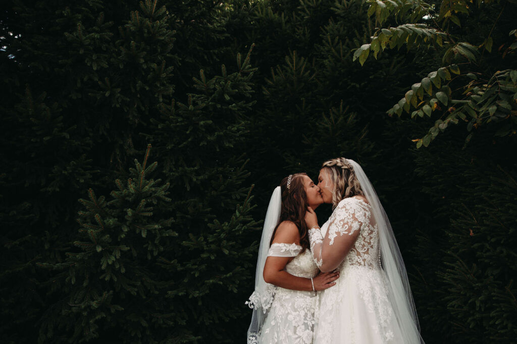 Brides kiss in front of green forest trees at greenhouse summer wedding.