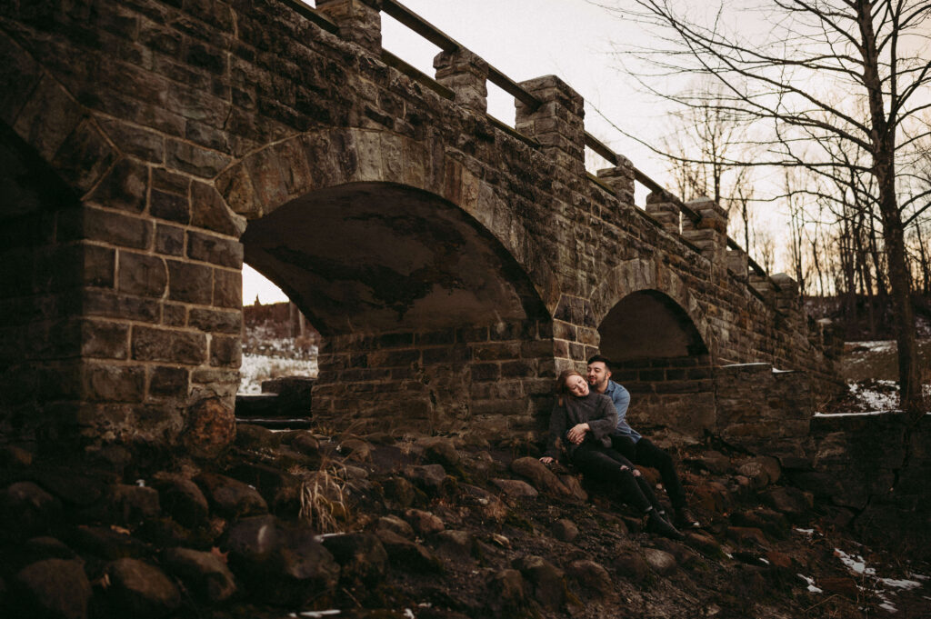 Engaged pair cuddle beneath a rock bridge similar to oens in Scotland.