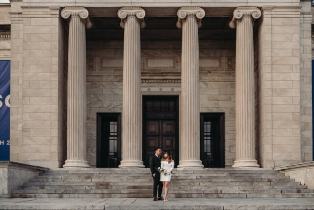 Married couple stand outside of Cleveland's museum in modern wedding clothes.